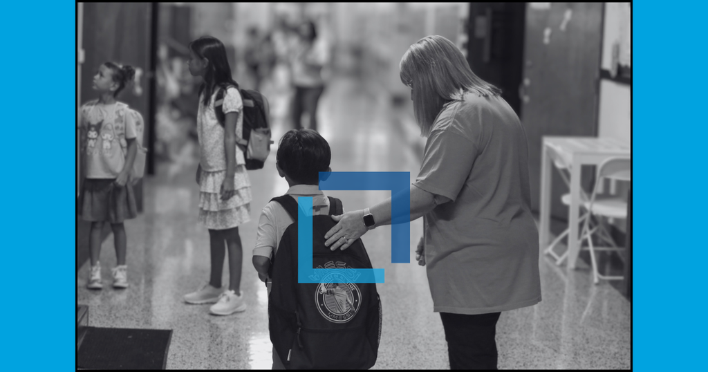 A school hallway with a teacher assisting a child with a backpack. Other children are standing in the background.