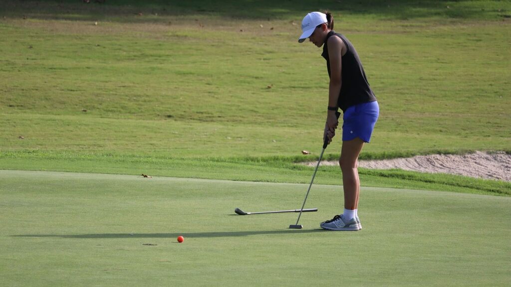 A golfer prepares to putt, standing on a golf course with green grass and a sand bunker.