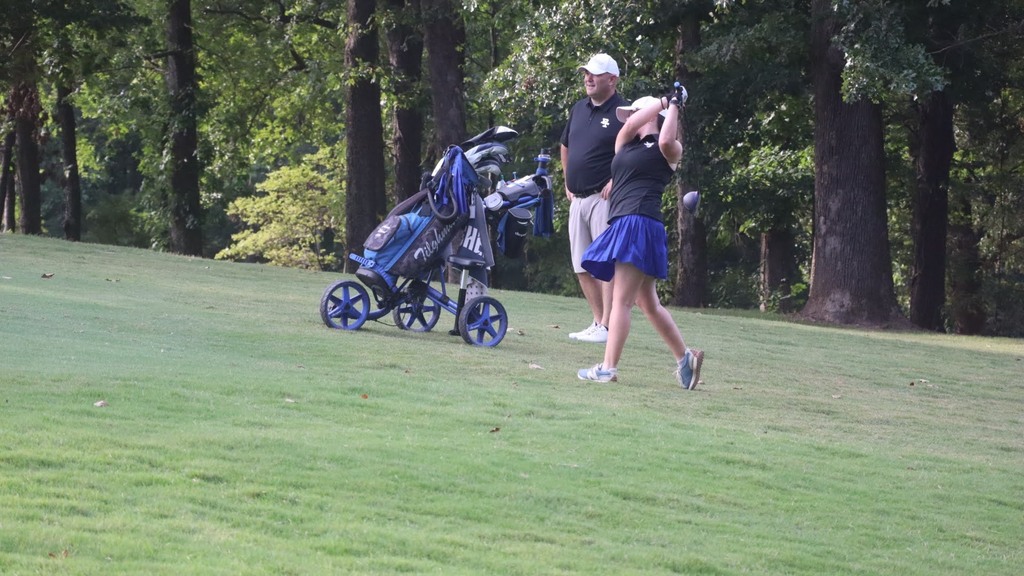 Person in skirt swings golf club on a grassy field. A golf cart stands nearby. Trees in background.