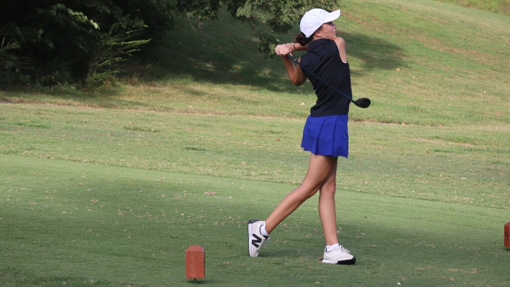 A person in a white hat and blue skirt swings a golf club on a grassy field.
