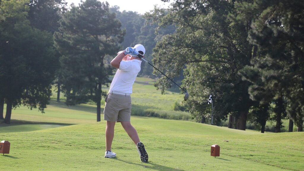 A golfer swings on a golf course, surrounded by trees and green grass. The golfer wears a white shirt, shorts, and a white cap.