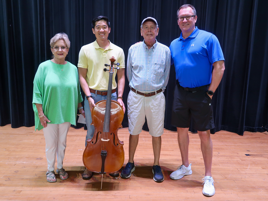 Four people stand on a stage with a cello, a black curtain backdrop, and wooden floor.