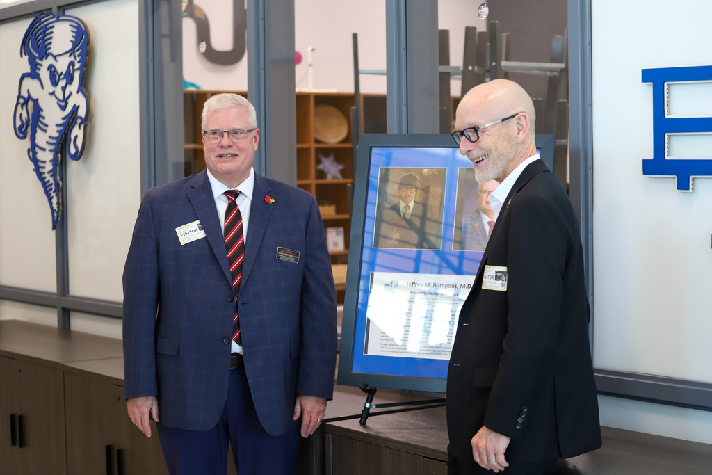 Two men stand before a poster; one wears a suit and tie, and the other wears glasses. The poster shows a photograph.