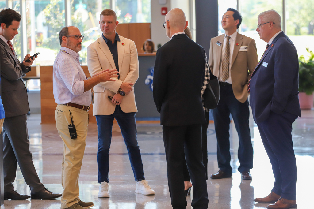 Men in suits and ties stand in a hallway. Some are talking. A potted plant is in the background.