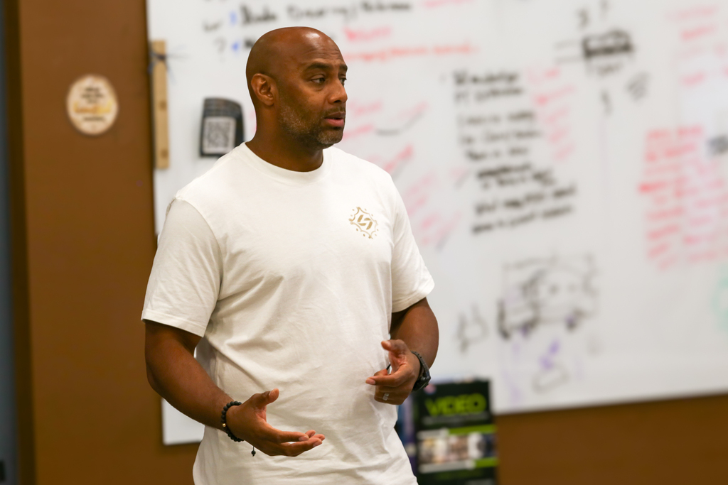 An adult male in a white T-shirt stands in front of a whiteboard covered in notes.
