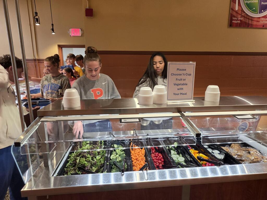 A buffet with various salads, vegetables, and other foods. A few people are standing behind the counter.