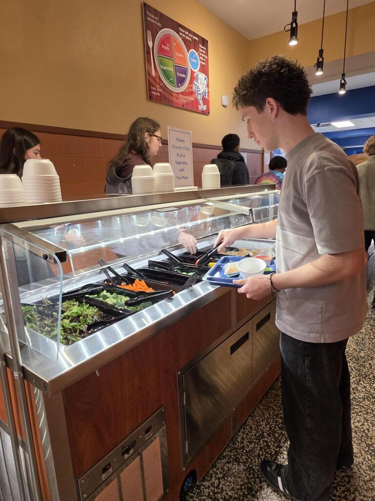 Man in gray shirt standing at a salad bar, picking up food, with people behind him.