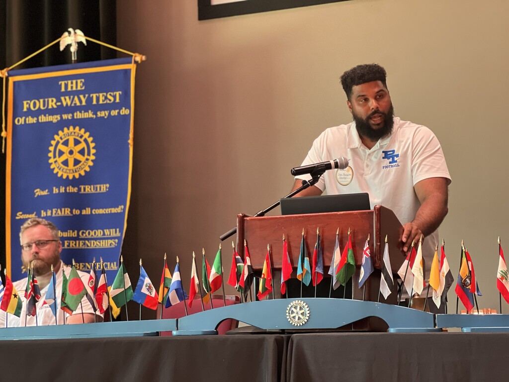 A person speaks at a podium with flags behind them. A banner on the left says "The Four-Way Test."