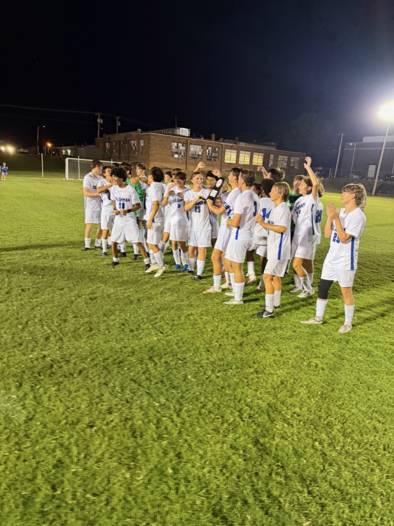 A group of young athletes in white uniforms, celebrating a victory on a grassy field at night.
