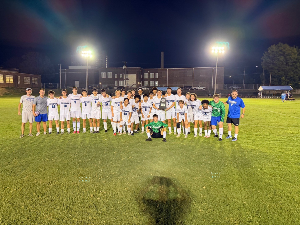 A soccer team poses on a field at night, wearing white jerseys and blue shorts. Lights and buildings are in the background.