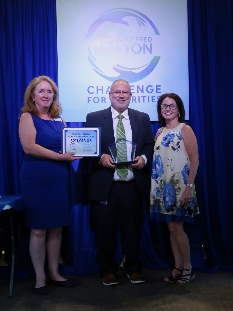 Three people stand in front of a blue curtain with a sign. A man holds an award. A woman holds a plaque.