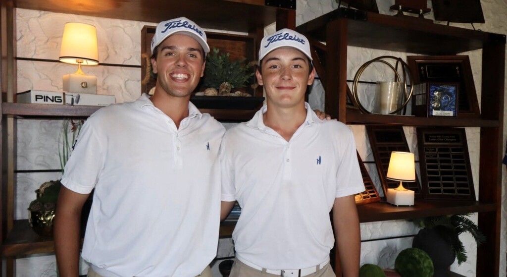 Two men in white polo shirts and caps, standing side by side in front of shelves with objects and plants.
