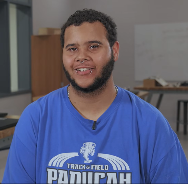 Man in a blue shirt with "TRACK & FIELD PANUCAH" logo, standing in a room with a wooden cabinet.