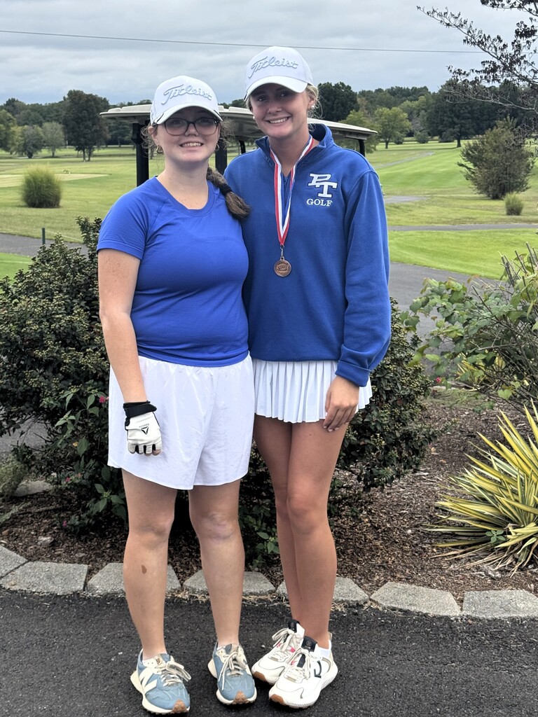 Two women in blue shirts and white shorts, one with a medal, stand on a path. Green bushes and trees surround them, with a golf course in the background.