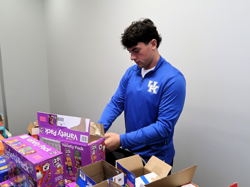 Man in a blue shirt opening purple box with boxes of food in front of a white wall.