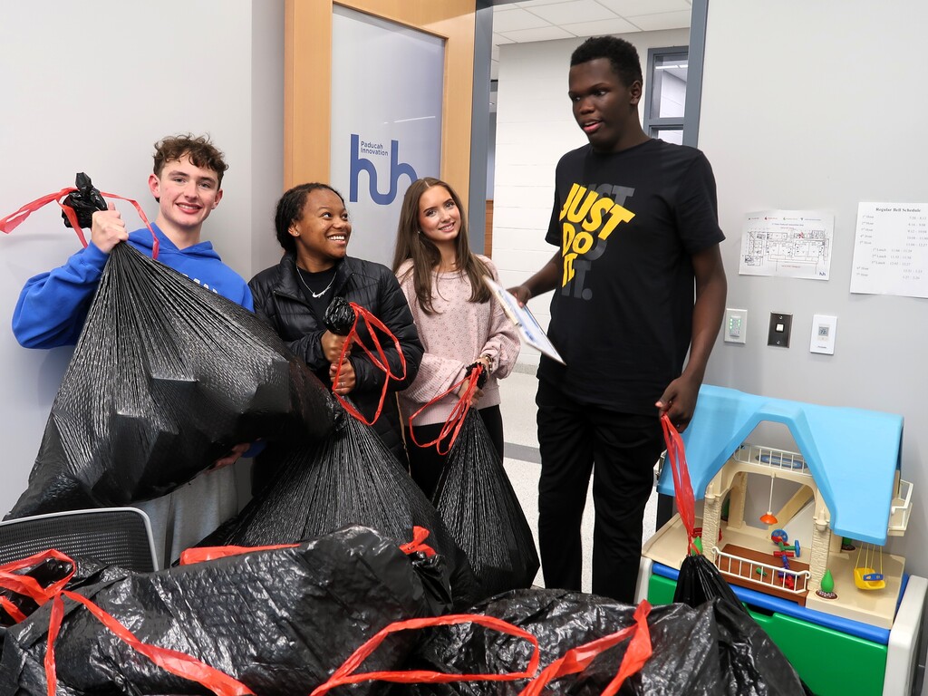 Four individuals hold bags of trash near a blue house model, one person holds a sign.