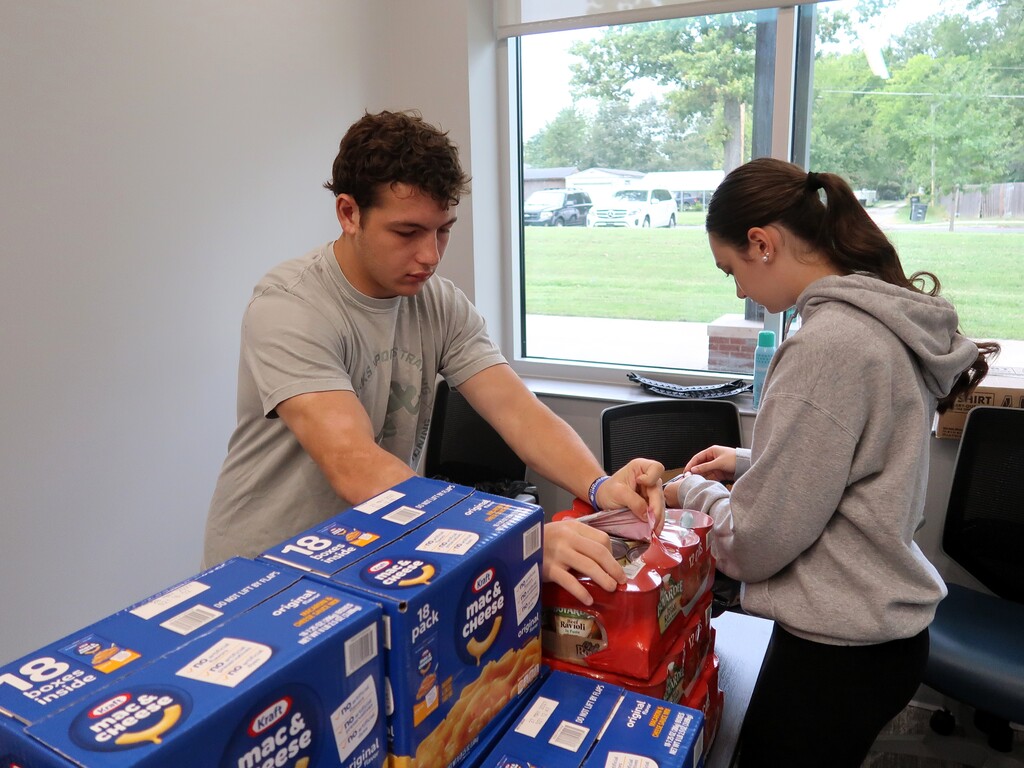 A man and a woman are packing boxes on a table. The boxes have a red logo. Behind them, a window overlooks a grassy field and trees.