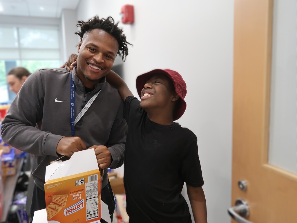 Two individuals in a hallway with one holding a cereal box, both smiling at the camera.