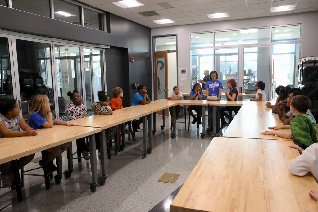 Children sit at a table with adults in a brightly lit room with large windows and gray walls.