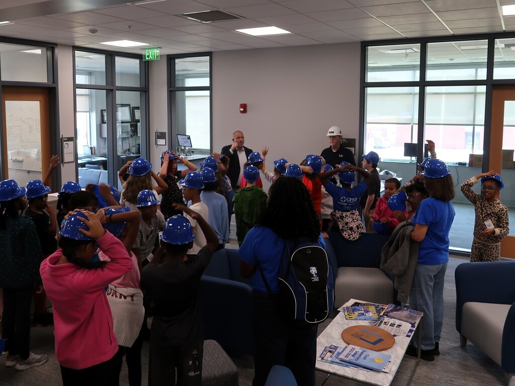 A group of people wearing blue helmets gather in a room with glass walls, standing and sitting.