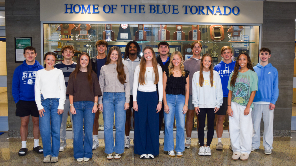 A group of teens stands in front of a glass display case inside a school.