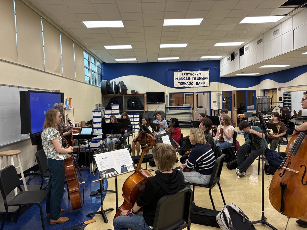 People sitting and standing in a music room, some holding cellos, facing a woman standing in front of a screen.