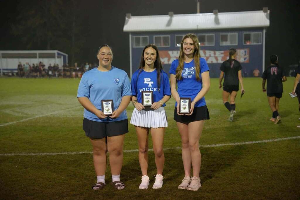 Three girls on a soccer field at night. Each holds a trophy and stands together, with two more players in the background.