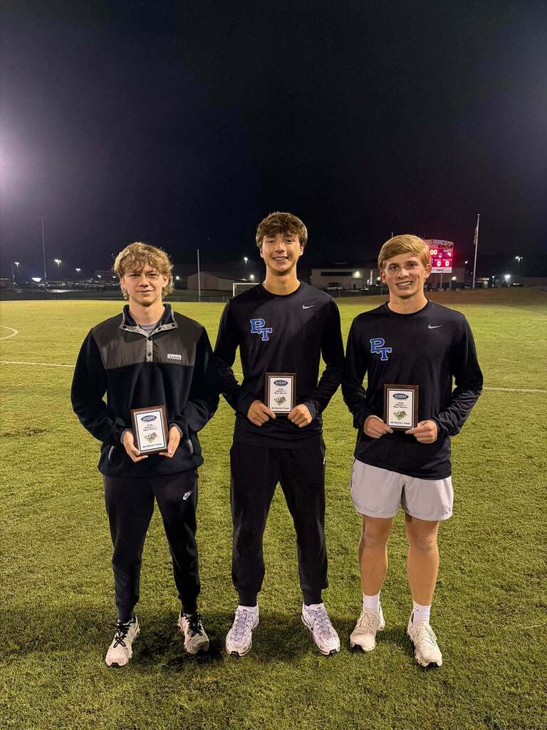 Three young men stand on a grassy field holding awards, two wearing black long-sleeve shirts, one wearing white shorts.