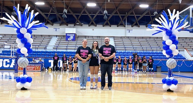 Three people in black t-shirts stand on a basketball court with blue and white balloon decorations.