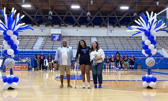 Three people stand on a basketball court, one using a cane. They are surrounded by blue and white balloons.