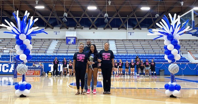 Four people in black T-shirts stand in a gymnasium with balloons. A woman holds a trophy.