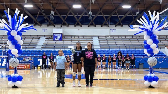 Three people stand in front of two balloon displays on a basketball court, with a team in the background.