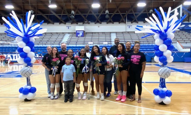 Group of people on a basketball court holding flowers and balloons, one person holds a volleyball.