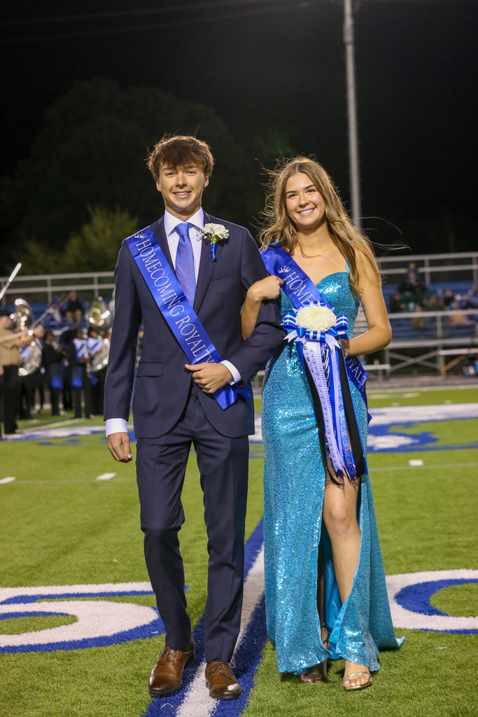 Teen couple wearing blue sashes walk on football field, band in background.