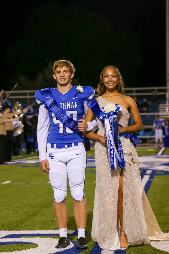 Two students, a young man in a football jersey and a woman in a long gown, pose for a photo on a football field.