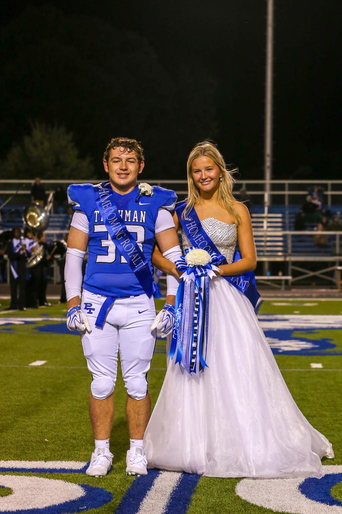 Man and woman wearing school colors, standing on football field, band playing in background, stadium in background.