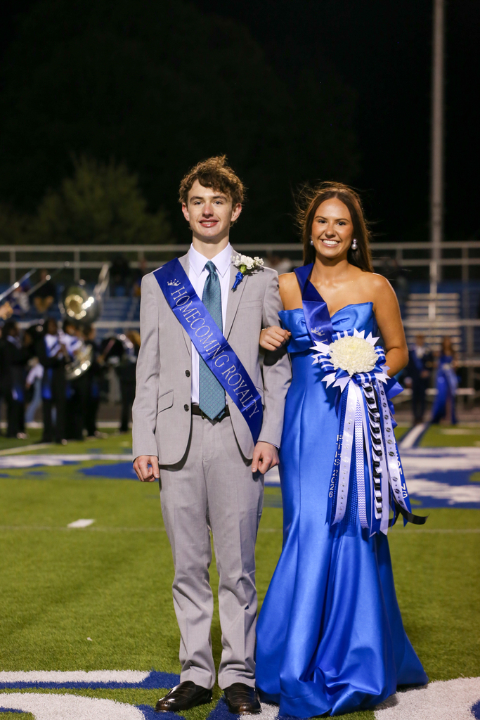 Teen boy and girl standing together on field, smiling. Boy has sash across chest, girl has sash over shoulder.