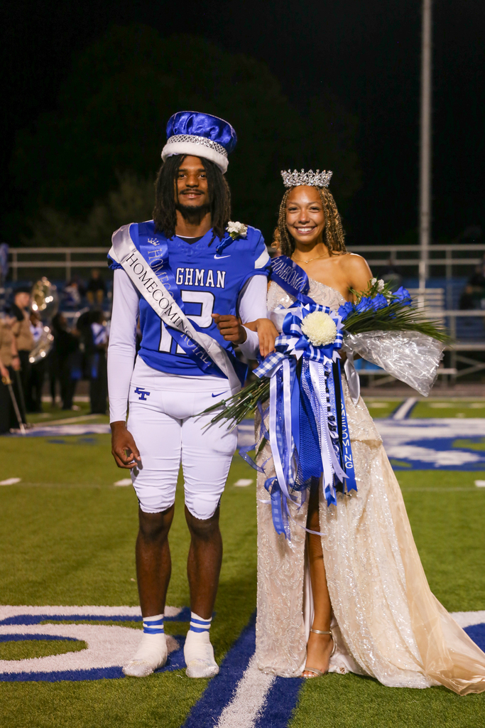 A young man and woman stand on a football field, both wearing crowns. The man is in a football uniform.