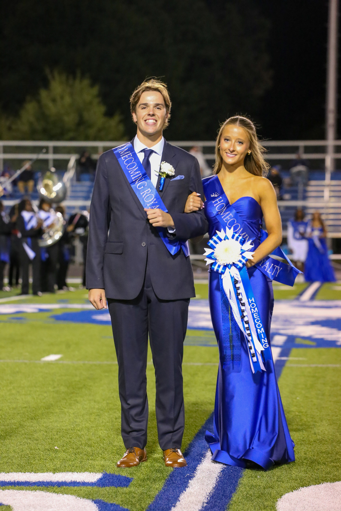 Two students dressed in formal attire stand on a football field. One holds a blue sash and the other holds a bouquet.
