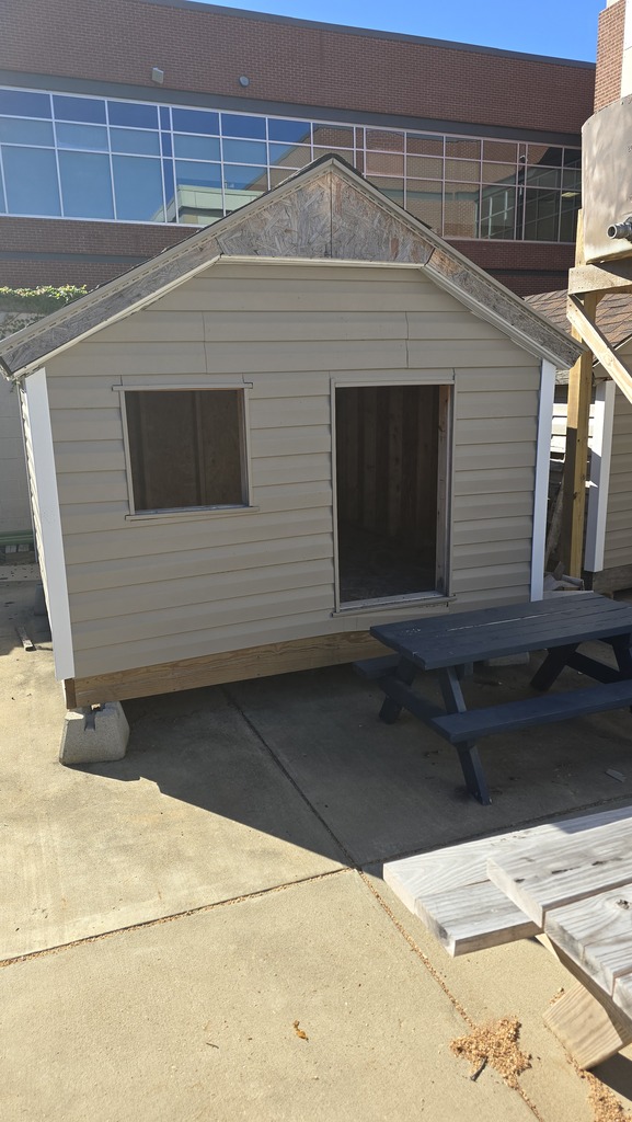 A small, light gray house sits on a concrete slab outside a building. It has two small windows and a large open door.