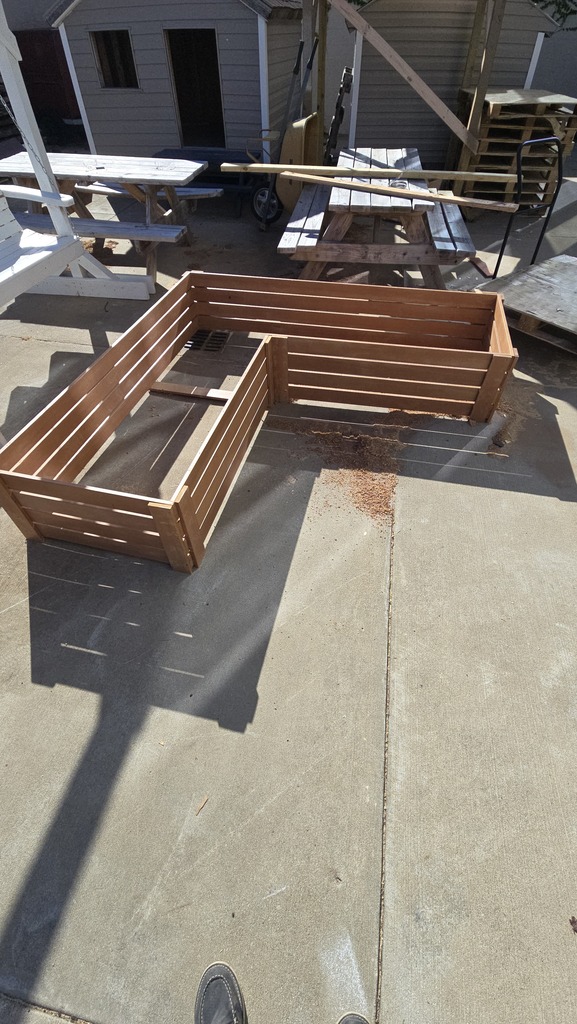 A wooden planter box on a patio with a concrete floor, near picnic tables and a building.