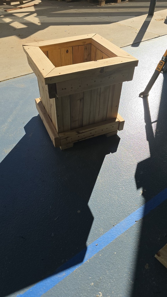 Wooden planter with vertical slats on a concrete floor with blue line. A shadow is cast.