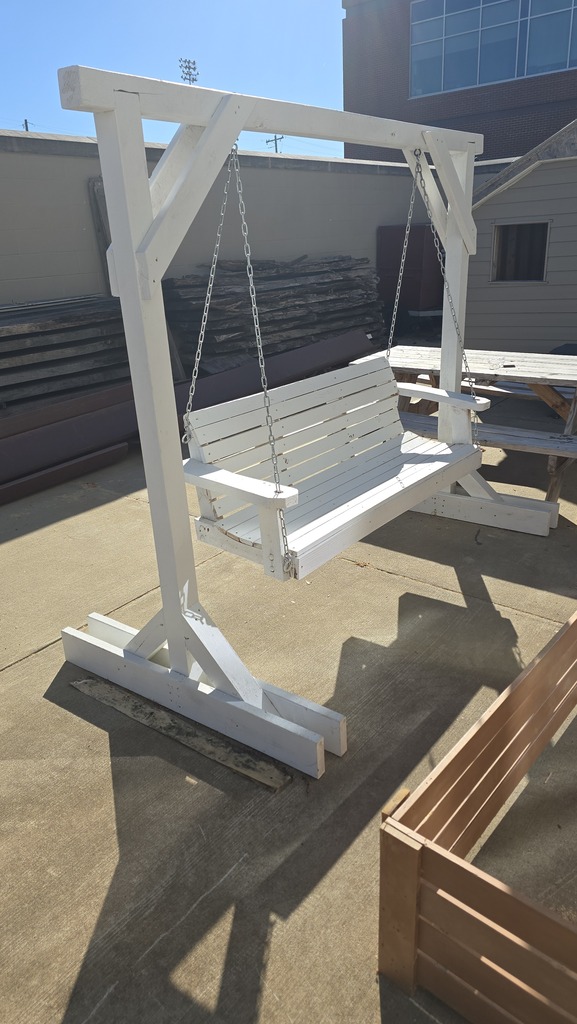 White bench swing on concrete with a wooden bench on right and a picnic table behind.