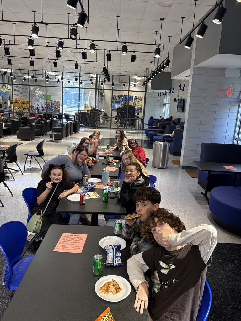 Students seated at tables in a cafeteria. Some eat pizza and one poses with an arm behind their head.