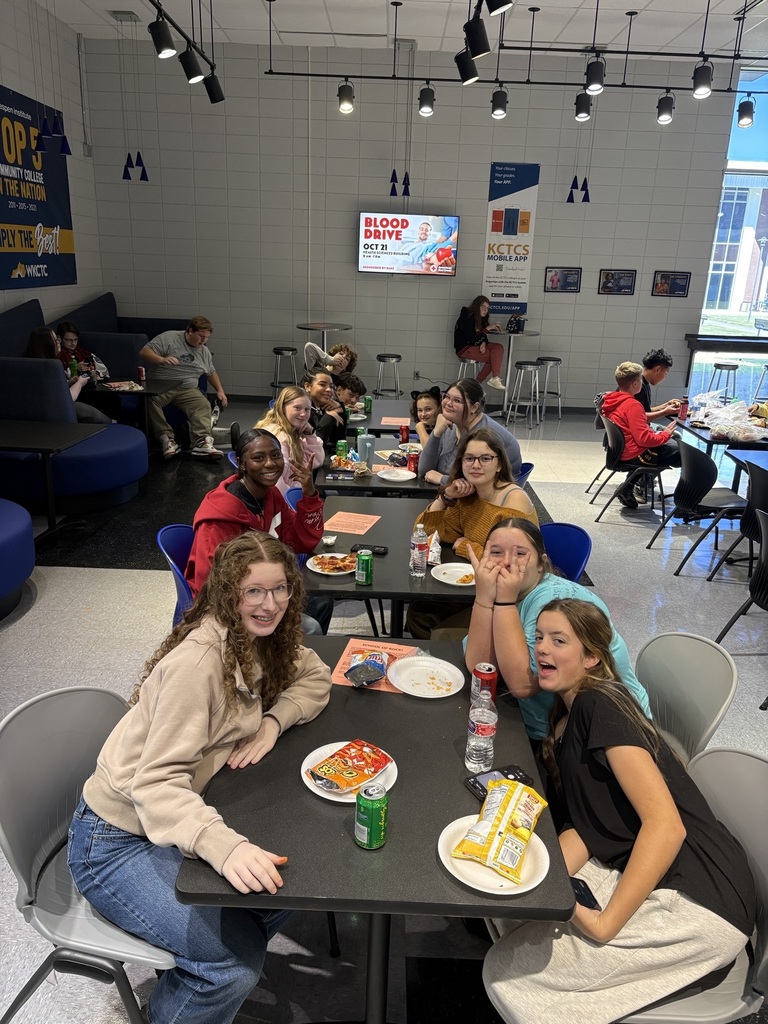 Students seated at tables in a common area. They are eating and talking. Plates, cups, and food on tables.