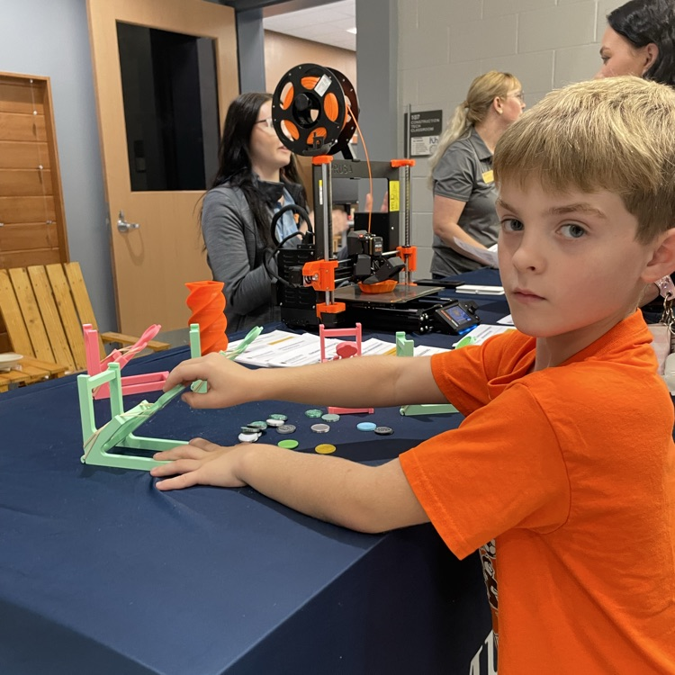A young boy is at a table, focusing on a toy setup while another child and adults observe.