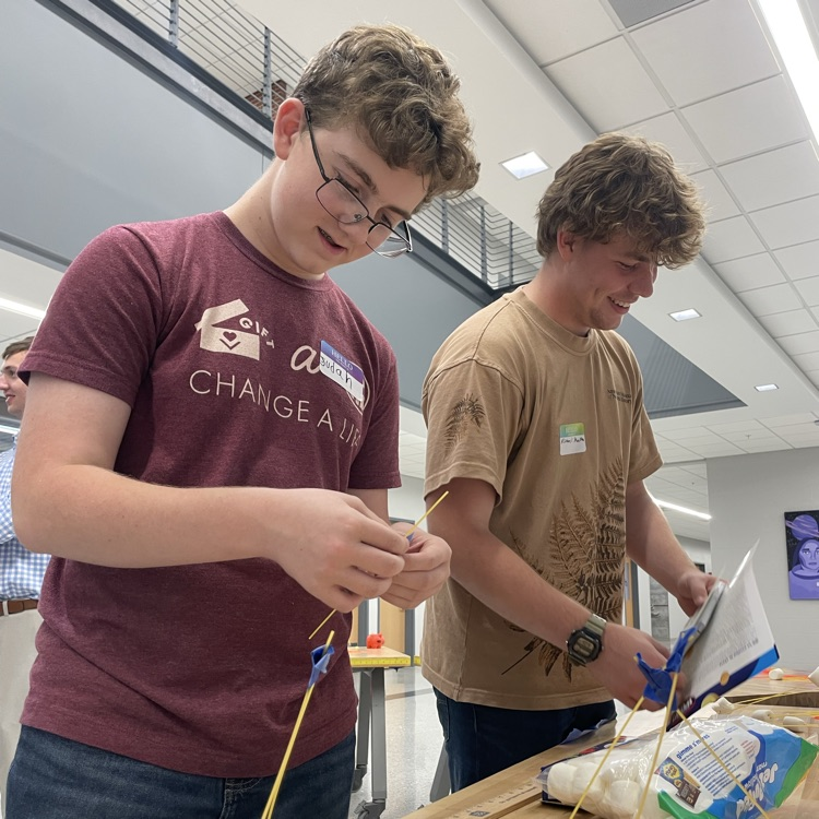 Two students working on a project with blue strings and a pile of items on a table.