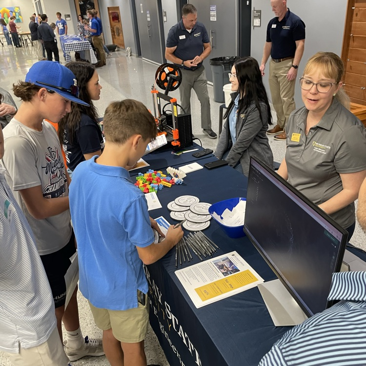 Students and adults gathered at a table with a computer and other items. Some people are standing.