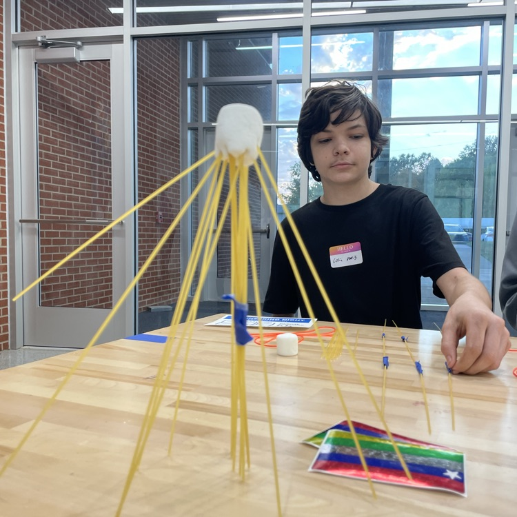 A student constructs a structure with yellow straws on a table. A colorful flag is nearby.