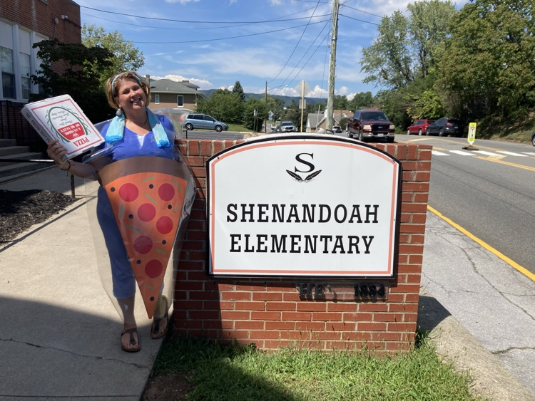 A smiling Shenandoah Elementary staff member dressed as a large pepperoni pizza slice stands beside the school sign while holding a pizza box to promote the Mamma Mia’s fundraiser.